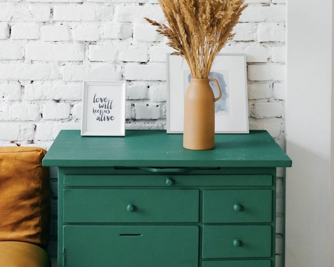 Green dresser with a brown vase of dried
          grass and framed art against a white brick wall
